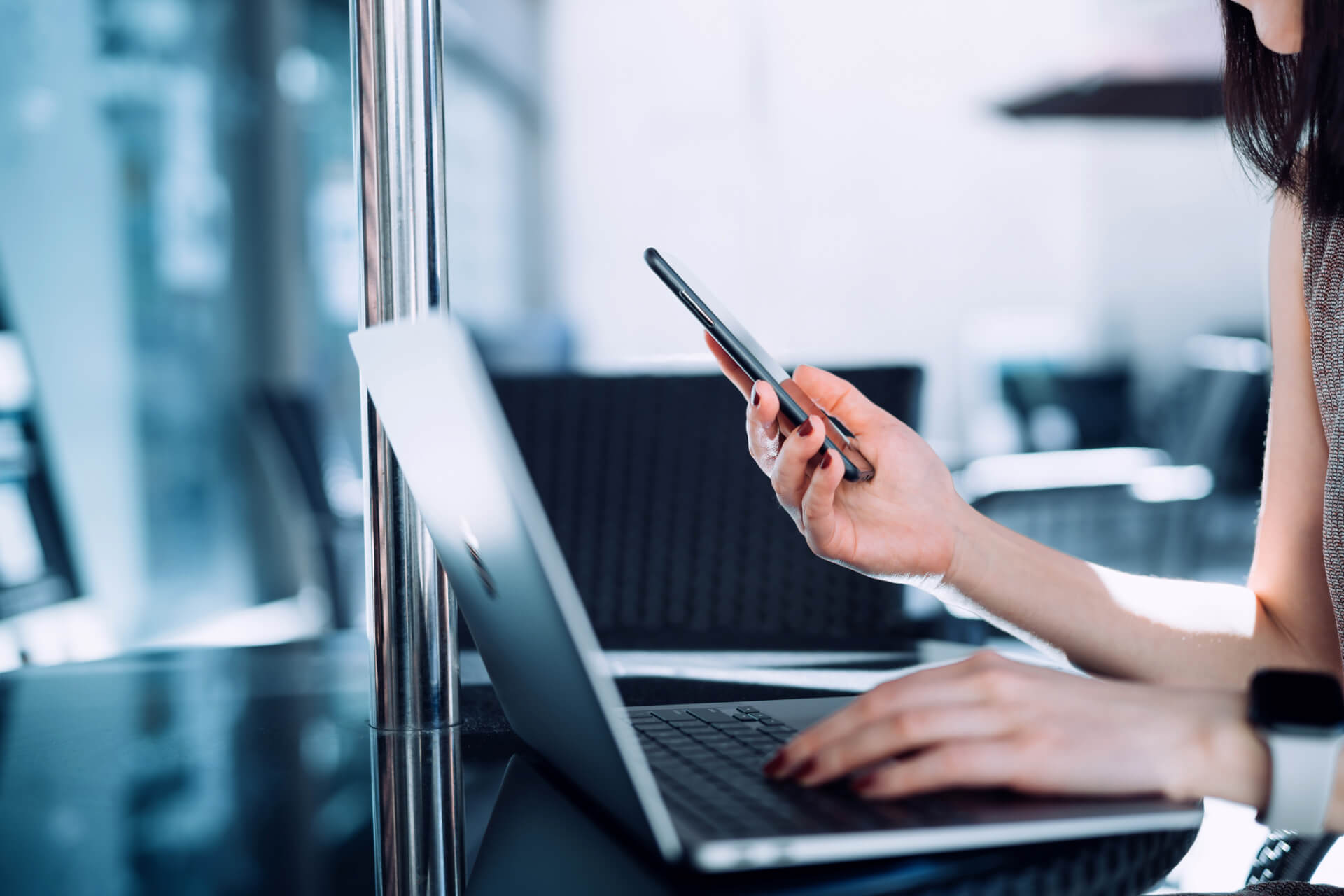 Cropped shot of young Asian businesswoman using smartphone and typing on keyboard on laptop on the go in sidewalk cafe. Remote working concept with flexible lifestyle.